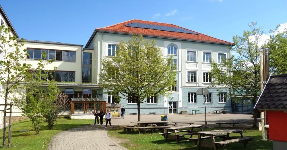 Außenaufnahme eines Schulgebäudes bei sonnigem Wetter. Im Vordergrund verläuft ein gepflasterter Weg durch eine grüne Rasenfläche mit Bäumen und Bänken.