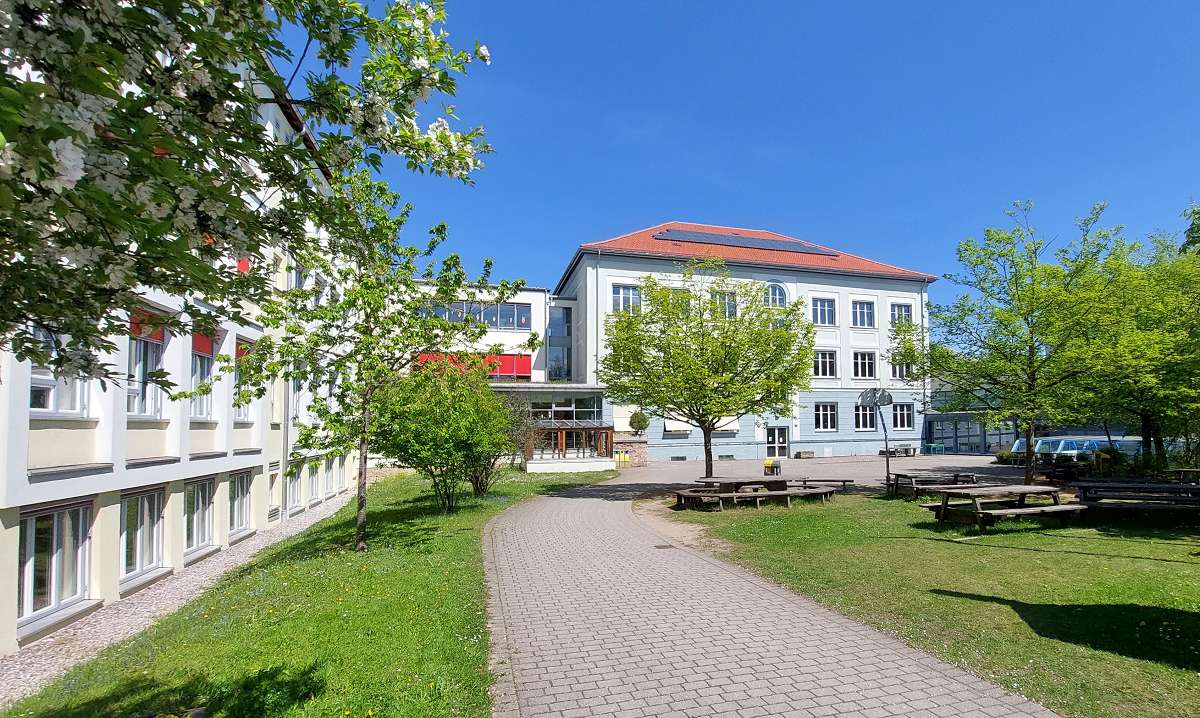 Außenaufnahme eines Schulgebäudes bei sonnigem Wetter. Im Vordergrund verläuft ein gepflasterter Weg durch eine grüne Rasenfläche mit Bäumen und Bänken.